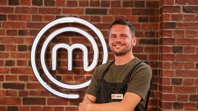 A man wearing a khaki t-shirt and an apron stood in front of the masterchef logo