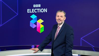 Gary Robertson in a blue suit standing in front of the words BBC Election 26