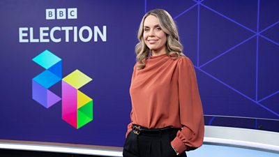 Laura Goodwin wearing an orange top and black trousers standing in front of the words BBC Election 26