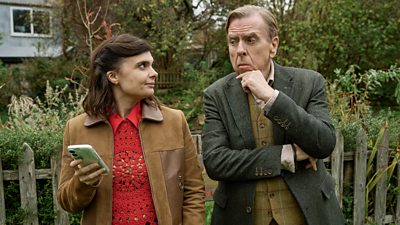 A man and woman stand side by side at a fence, looking to each other. She holds a mobile phone, he holds a hand to his chin.