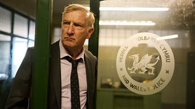 A man in a suit and tie looks out the door of an office. Mid Wales Police is written in English and Welsh on the glass.