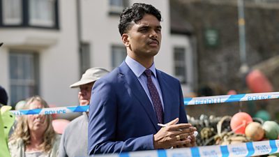 A man in a suit stands behind police tape taking notes