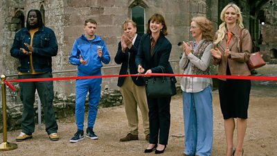 A group of people gather together outside a building, standing behind a red ribbon that's being cut ceremonially