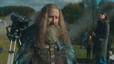 Jim Howick in character as Randall St.Clair. He’s dressed in a historical costume and wears a long wig, a beard, and a green cloak over a tunic. A TV camera on a tripod is visible in the background. 