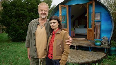 Owen Teale and Gwyneth Keyworth in character as Micheal Mallowan and DS Janie Mallowan respectively. Micheal smiles and has his arm around Janie. Janie looks uncertain. A small lodge is in the background, the doors are open and a bedroom is visible inside. 