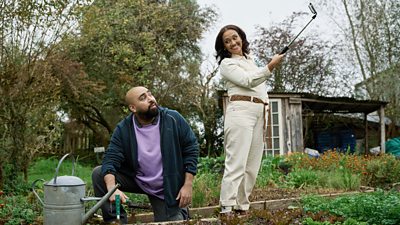 Asim Chaudhry and Tori Allen Martin in character as Omar and Amanda respectively. They are both outside in a garden. Omar kneels with a trowel to tend to flowerbeds, while Amanda grins and holds a gardening tool aloft.  