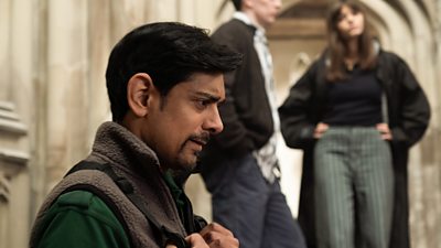 Amit Shah in character as Ash. He appears in an old, stone building. Sat in the foreground of the image, two other characters are blurred in the background. 