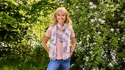 Michaela Strachan wearing a pink shirt, scarf and blue jeans smiling towards camera. She is standing in a woodland surrounded by greenery