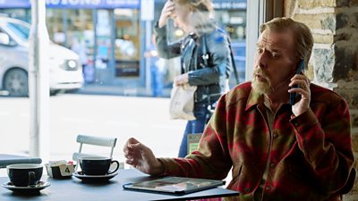 A man wearing a brown and red jacket. He's sat in a cafe with a mug in front of him while he's on the phone