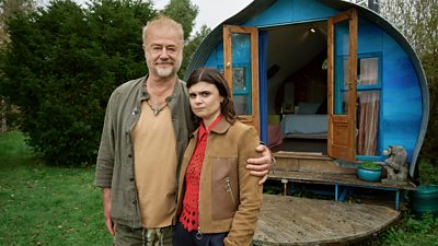 A middle-aged man wearing a green shirt with his arm around a younger women wearing a beige jacket. They both look towards the camera standing in front of a small lodge
