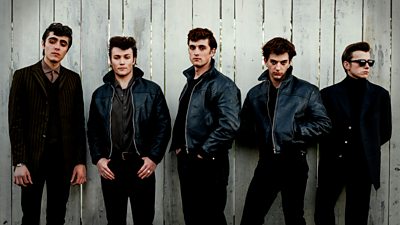 Five men lined up against a white fence with four looking to camera. All the men are wearing dark clothes with three wearing leather jackets