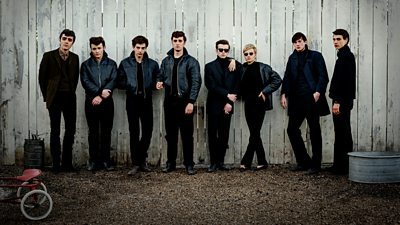 Eight people all wearing black clothing against a white picket fence, all of them have a stern look as they stare into the camera