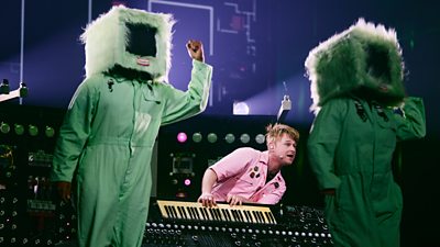 A man in a pink jumpsuit playing a keyboard. To either side are two figures in green jumpsuits with fluffy computers for heads