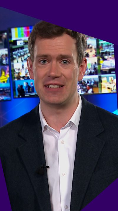 Phil Sim - man in shirt and suit, standing in front of a bank of TV monitors