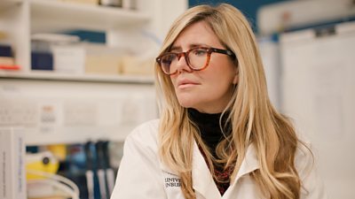 A woman in a scientific lab coat sits in a laboratory and watches something offscreen.
