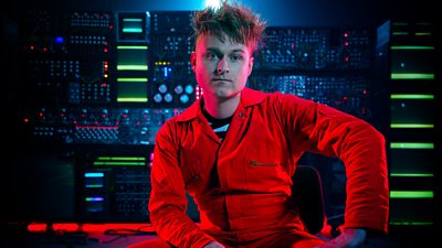 A man in a red boiler suit sits in front of a large machine with glowing green lights