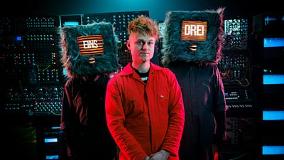A man in a red boiler suit stands in front of two figures in boiler suits with furry computer monitors on their heads. The screens read Eins and Drei. The three stand in front of a large series of machines.