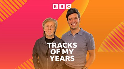 Sir Paul McCartney and Vernon Kay stand side by side in a room, smiling to camera. Paul wears a denim style jacket over a black t-shirt and Vernon wears a blue polo shirt