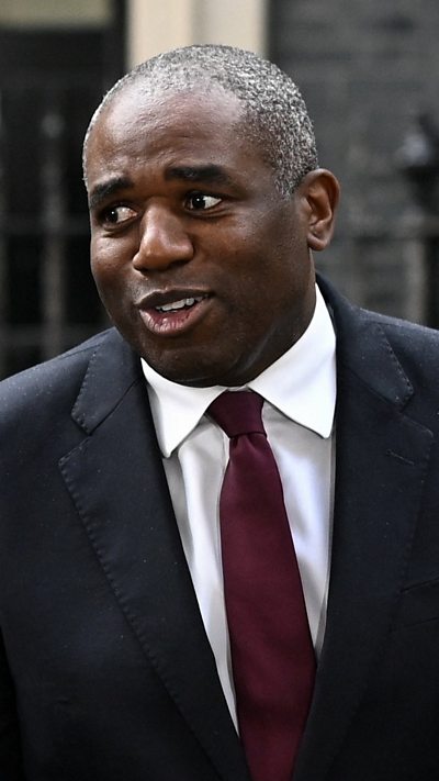 A mid-shot of David Lammy wearing a black suit with white shirt and maroon tie looking off-camera.