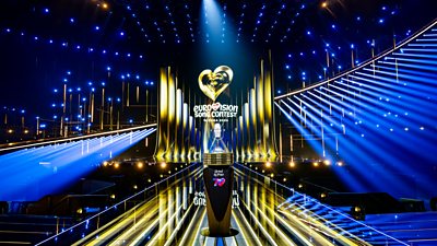 The glass microphone Eurovision trophy sits on a plinth on stage in Vienna, with beans of yellow and blue light shining around it