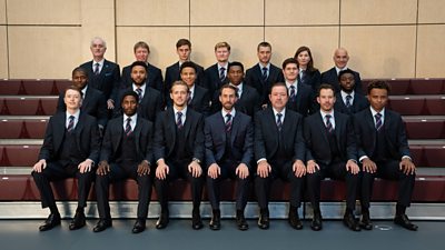 A row of three footballers sat down for a team photo. They are wearing matching navy suits, blue shirts and striped ties