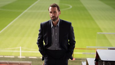 A man wearing a smart suit looking into the camera while stood in the seating area of a football stadium