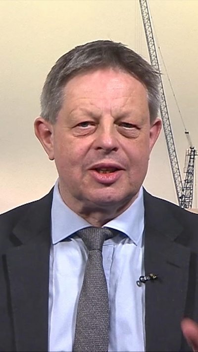Iain Watson wearing dark suit, light coloured blue short and a grey tie