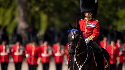 A soldier in ceremonial uniform rides a horse in central London. Other soldiers in red jackets and fur hats are visible in the background.