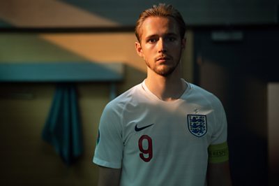 A man wearing a white England number 9 shirt, he is looking directing into camera stood in a dimly lit locker room