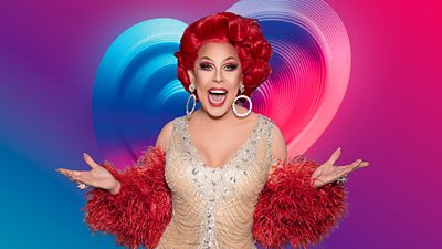 La Voix, a drag performer in a glittering dress with a bright red wig, smiles to camera, set against a backdrop of a pulsing blue, pink and purple heart