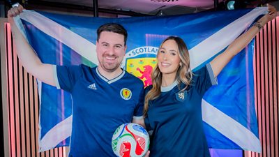 Steven Mill and Amy Irons wearing Scotland football shirts and holding up a Scotland football flag together 