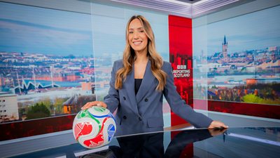 Amy Irons wearing a grey blazer, holding a football, stood in a news studio and smiling towards camera
