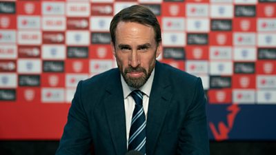 A man in a suit sits at a press conference looking straight down the camera