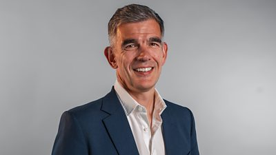 A man in a navy blue suit jacket and white short smiles to camera, set against a grey background