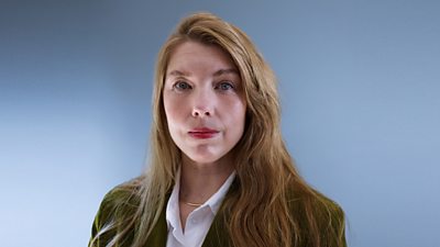 A woman in a green blazer and white shirt stands against a grey background.