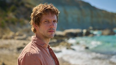 Ο Damien Molony στον χαρακτήρα του Dan. Στεκόταν σε μια αμμώδη παραλία στον ήλιο, φοράει ένα ανοιχτό πουκάμισο και παρακολουθεί κάτι εκτός οθόνης. 