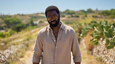 Ο Nicholas Pinnock στον χαρακτήρα του Solomon. Φορώντας γυαλιά ηλίου και πουκάμισο, περπατά σε έναν δρόμο κάτω από τον ήλιο. Μια σειρά από κάκτους πλαισιώνουν την άκρη του δρόμου. 