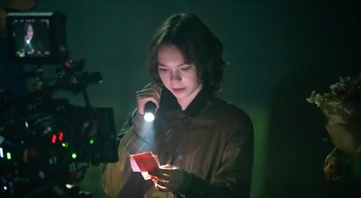 A girl shines a torch light on some sheets of paper as a camera crew films her