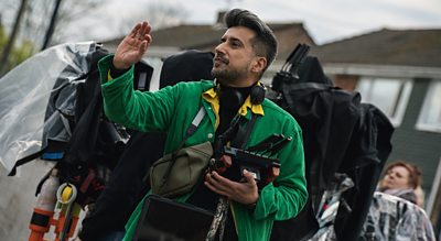 A man stands outdoors on set in a green jacket, directing