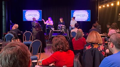 Panel discussion on stage with four speakers and two presentation screens, watched by a seated audience.