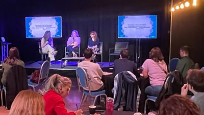 Panel discussion with three speakers on stage and two presentation screens, viewed by a seated audience.