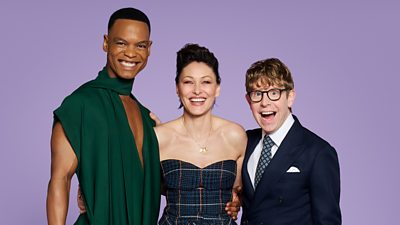 Three people stand smiling to camera. Johannes wears a long green jumpsuit, Emma wears a tartan corset and Josh wears a suit and tie