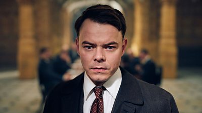 Charlie Heaton as Charles Shelby in Peaky Blinders. Wearing a suit and tie, he stands in the foreground. Blurred in the background are people sitting at a long conference table.