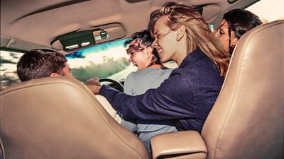 Four people in a car. The photo is taken from the backseat, three people on the right of the car, look to the driver on the left. 