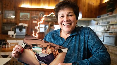 A woman smiles to the camera, while holding a photograph. 