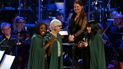 Claudia Winkleman and Traitors cast members, Minah Shannon and Linda Rands, all wear green long cloaks and laugh, standing in front of an orchestra.