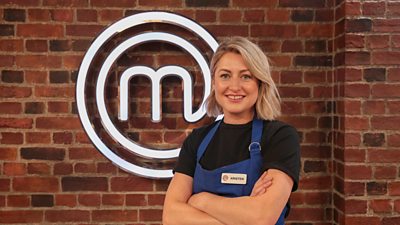 A woman wearing a black t-shirt and blue apron stood in front of a glowing masterchef logo