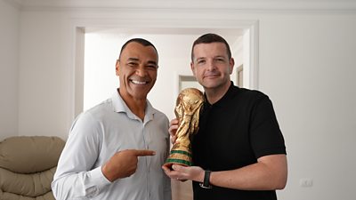 Kevin Bridges wearing a black polo, smiling towards camera as he holds the world cup trophy next to ex-footballer Cafu