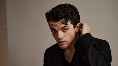 A head and shoulders shot of a man with dark hair in a dark jacket, looking to camera with one hand reaching around to the back of his neck