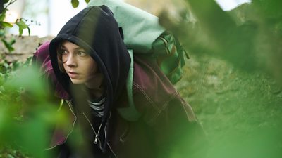 Pip wearing a hoodie and a rugsack hiding in some bushes
